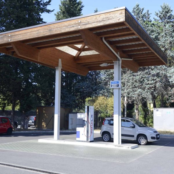 Punto di ricarica auto elettriche sotto tetto in legno in area verde con veicoli e alberi.